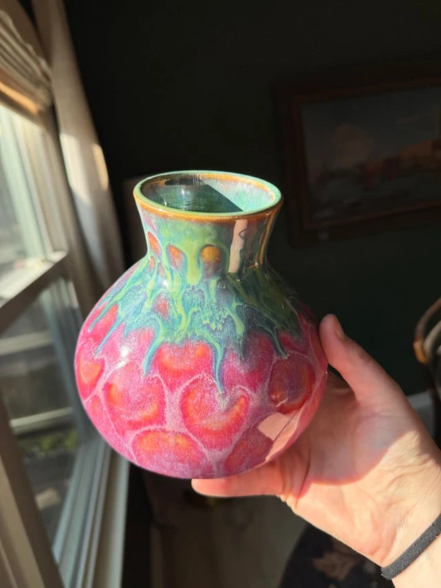 A pink ceramic vase with green and yellow glaze, held in a hand beside a window