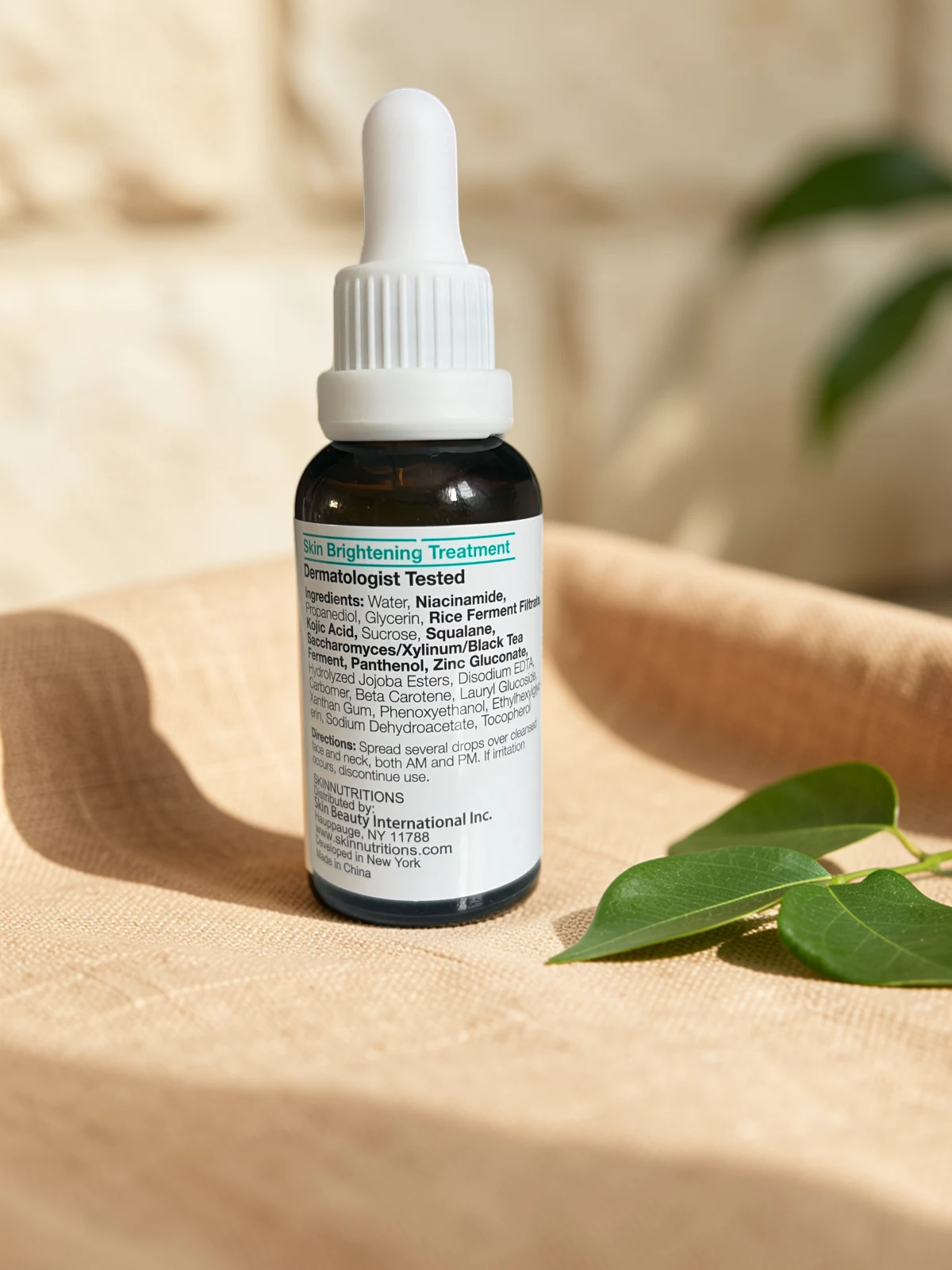 The same serum bottle on sand-colored linen beside a green leaf, in soft window light